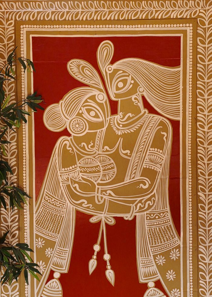 Intricate Indian folk art depicting a couple in vibrant earthy tones on a rich red backdrop.